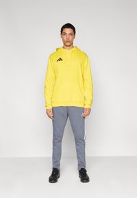 Κίτρινο φούτερ με κουκούλα και μπροστινή τσέπη με μαύρο λογότυπο· συνδυάζεται με γκρι φόρμα με λευκές πλευρικές ρίγες και μαύρα αθλητικά παπούτσια.