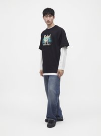 Νέος άνδρας που φοράει μαύρο γραφικό t-shirt πάνω από λευκά μακριά μανίκια, μπλε τζιν και μαύρα αθλητικά παπούτσια, στέκεται μπροστά σε λευκό φόντο.