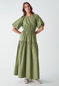 Abito maxi verde oliva con maniche a sbuffo, vita elastica, strati arricciati e texture morbida. Abbinato a scarpe slip-on in tessuto bianco.