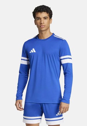 Junger Mann trägt blaues langärmeliges Sporttrikot und passende Shorts mit weißen Streifen, steht vor einfachem Hintergrund.