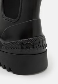 Botte en caoutchouc noir avec des accents texturés, dotée d'un logo TOMMY proéminent et d'une semelle striée pour une meilleure adhérence. Surface lisse avec finition mate.