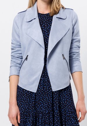 Frau trägt eine hellblaue Jacke mit Reißverschluss über einem marineblauen Kleid mit kleinen blauen und weißen abstrakten Flecken.