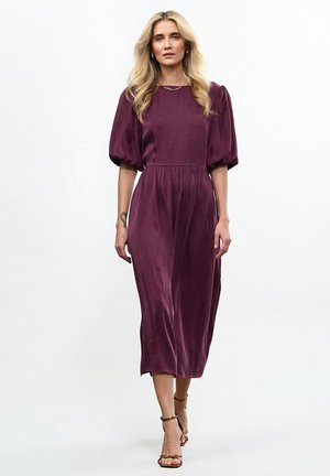 Fioletowa sukienka midi z bufiastymi rękawami i dopasowaną talią, wykonana z miękkiego, teksturowanego materiału, zestawiona z brązowymi sandałami na paski.