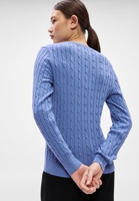 Gaiši zils džemperis ar pinuma rakstu, ar ribotiem aprocēm un apakšu, teksturētu rakstu, kas vertikāli izkrāsots gar ķermeni.