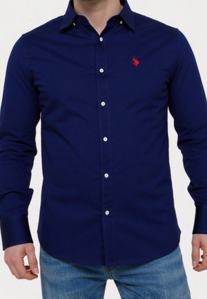 Man draagt een nauwsluitend, lang mouw navyblauw overhemd met kleine rode geborduurde logo op de borst, gecombineerd met lichtblauwe spijkerbroek.