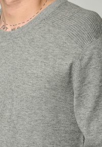 Pull gris en textile doux, avec des accents côtelés aux épaules et un col rond, présentant une finition lisse et des variations de texture subtiles.