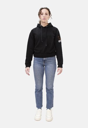 Donna in piedi, rivolta in avanti, che indossa una felpa nera con cappuccio e logo sulla manica, jeans blu e scarpe da ginnastica bianche su uno sfondo semplice.