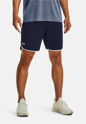 Man draagt marineblauwe Under Armour sportshorts, lichtgrijze sportschoenen en een blauw patroon overhemd, staande op een witte achtergrond.