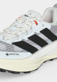 Λευκό και γκρι παπούτσι τρεξίματος Gore-Tex με μαύρες ρίγες Adidas, λευκά κορδόνια με γκρι σχέδια και κρεμ χρώματος σόλα.