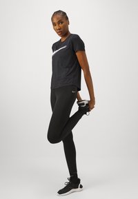 Must musta t-särk, millel on valge Nike logo, koos mustade legging'ute ja mustade spordijalatsitega. Mudel venitab ühte jalga.