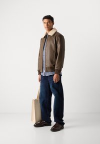 Giacca bomber in pelle marrone con colletto in shearling crema, camicia a quadri, jeans blu scuri e scarpe marrone scuro, accanto a una borsa tote beige.