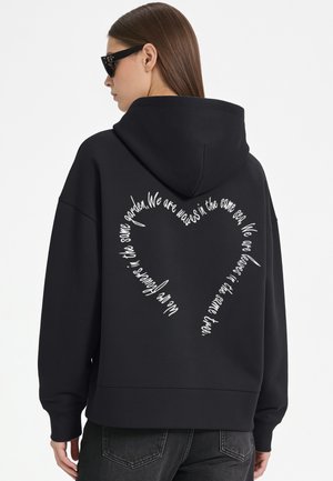 Femme portant un sweat à capuche noir avec un texte blanc en forme de cœur dans le dos, debout de profil sur un fond uni.