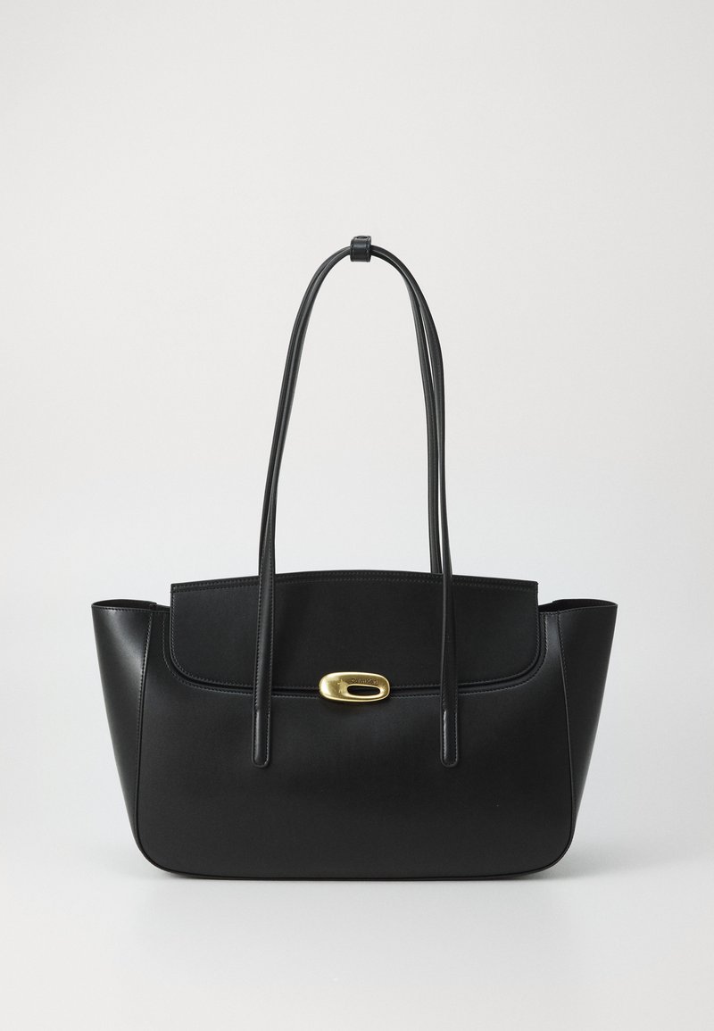 Sac en cuir noir à bandoulière avec de longues sangles et un fermoir ovale doré sur fond gris clair uni.