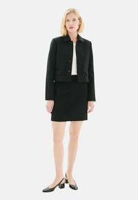 Veste noire cintrée avec un col, des boutons avant et une silhouette ajustée, associée à une jupe noire et des ballerines noires à bout pointu. Texture lisse.