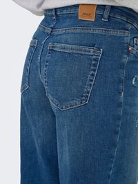 Blugi din denim albastru cu buzunare în spate, etichetă de marcă din piele maro deschis inscripționată cu „ONLY”, purtați cu un top gri deschis.