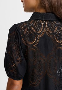 Maglietta nera con scollatura a colletto, caratterizzata da dettagli in pizzo intricate, maniche a sbuffo e un tessuto testurizzato.