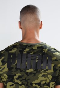 Grön kamouflage-t-shirt med en svart "PUMA"-logotyp på baksidan. Tillverkad av bomull, har rund halsringning och korta ärmar.