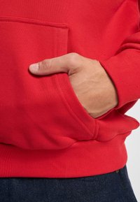 Felpa rossa con cappuccio, tasca a marsupio, polsini e orlo ribattuti. Il tessuto appare morbido e texturizzato, con una vestibilità comoda e un design informale.