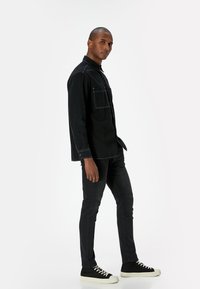 Chaqueta de denim negra con dos bolsillos frontales, costuras blancas y un corte relajado, combinada con pantalones ajustados negros y zapatillas negras con suelas blancas.