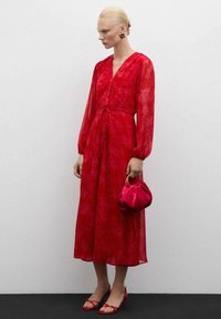 Rood bloempatronen jurk met lange, doorzichtige mouwen, een strakke taille en een midi-lengte zoom. Gecombineerd met een kleine rode handtas en bijpassende sandalen.