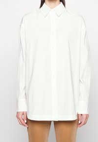 Vit skjorta med knappar i bomull. Har en klassisk krage, lång ärm med vertikala veck och en subtil logotyp på bröstet.