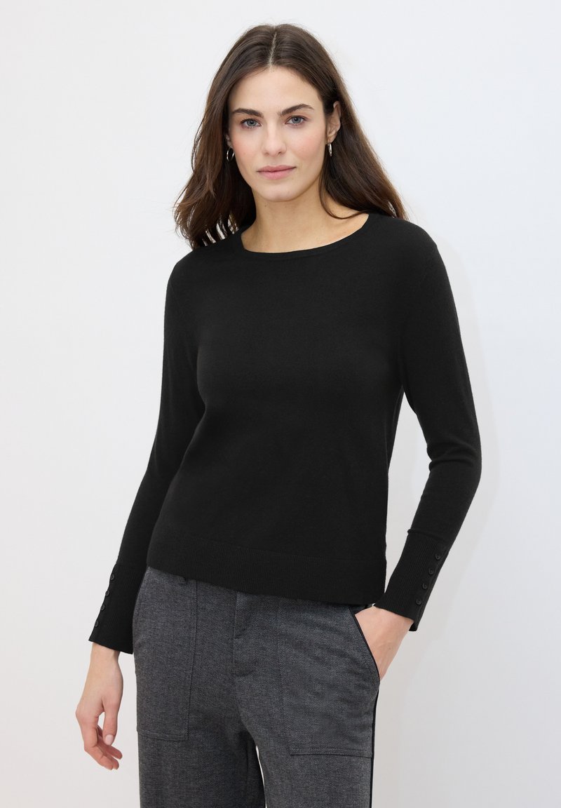 Pull en maille noire avec un col rond, des manches longues et des accents de boutons aux poignets, associé à un pantalon gris sur mesure. Texture lisse.