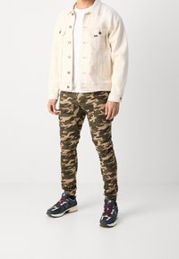 Lichtbeige denimjack, witte t-shirt, camouflage cargopantalon en multicolore sportschoenen met zwarte en marineblauwe accenten.