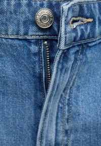 Gros plan sur la braguette d'un jean en denim bleu avec une fermeture éclair métallique partiellement ouverte, un bouton argenté portant l'inscription « MNG JEANS ÉTABLI EN 1984 » et une boutonnière cousue.