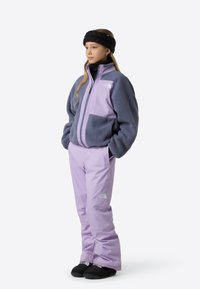 Vijolična flis jakna s sivimi poudarki, kombinirana z ujemajočimi vijoličnimi snežnimi hlačami. Črna traka za glavo in natikači dopolnjujejo obleko.