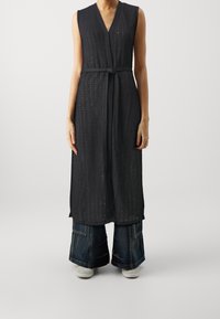 Donna che indossa un cardigan senza maniche in maglia scura con cintura, abbinato a jeans in denim scuri a gamba larga e sneakers bianche, contro uno sfondo neutro.