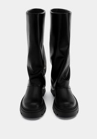 Botas altas de cuero negro con un diseño holgado, punta redondeada, suela de goma gruesa y detalles mínimos sobre un fondo liso.
