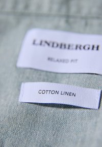 Tøjmærker på lysegrønt stof med mærket "Lindbergh", "Relaxed Fit" og materialet "Bomuld-linned".
