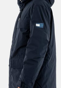 Giacca imbottita di colore blu navy con cappuccio, tasche laterali e un patch con logo sulla manica. Tessuto liscio e strap per polsini regolabili.