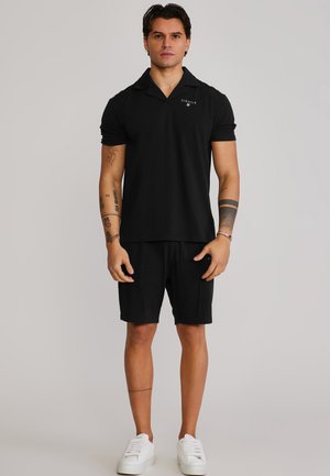 Ein Mann steht frontal und trägt ein schwarzes kurzärmeliges Hemd mit Kragen, schwarze Shorts, weiße Turnschuhe sowie sichtbare Tattoos an Armen und Beinen.