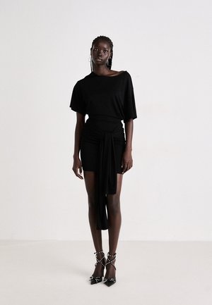 Abito nero con design corto e asimmetrico, con un dettaglio annodato in vita. Abbinato a tacchi alti neri con accenti borchiati.