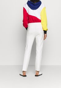 Bunte, gekürzte Kapuzenjacke mit blauen, roten, weißen und gelben Panels; weiße, maßgeschneiderte Hose; schwarze Schlippersandalen; und glatte Stofftextur.