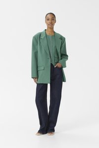 Overdimensjonert grønn blazer med skarpe slag, frontlomme, kombinert med tettsittende mørkeblå jeans og lyse sandaler, mot en hvit bakgrunn.