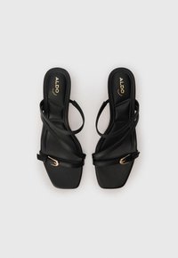 Black strappy sandals with a square toe, made of textured material. Features thin, overlapping straps and a small gold buckle detail.