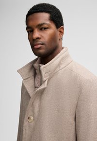 Man met kort krullend haar draagt een beige wollen jas over een lichtroze ritsjack, met een lichte draai naar links en een neutrale uitdrukking op een grijze achtergrond.