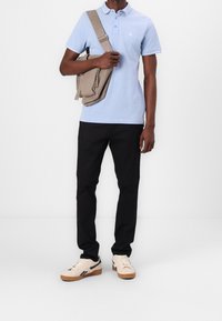Polo shirt bleu clair à manches courtes avec un col, pantalon noir et baskets beiges. Un sac à bandoulière gris est porté sur l'épaule.