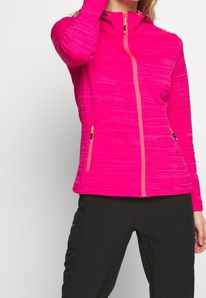 Vrouw draagt een felroze jack met ritssluiting en zijzakken, gecombineerd met een zwarte broek, poseert met één hand op haar hoofd.