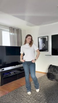 Valkoinen lyhythihainen t-paita, yhdistetty sinisiin farkkuihin ja valkoisiin kenkiin. Taustalla musta televisio ja taideteos seinällä.