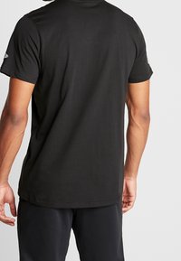 Hombre con camiseta negra de manga corta y pantalones negros, visto desde atrás contra un fondo claro y liso.
