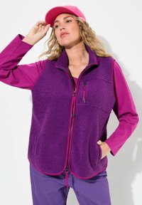 Lila Fleece-Weste mit Reißverschluss vorne, kontrastierenden pinken Akzenten, Seitentaschen und einem glatten langärmeligen Hemd darunter.