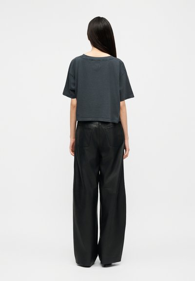 Hosszú, sötét hajú nő, laza, sötétszürke rövid ujjú felsőt és bő szárú fekete bőrnadrágot viselve, fehér háttér előtt háttal állva.