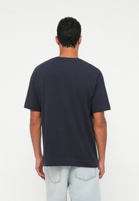 Μπλε βαμβακερό T-shirt με κοντά μανίκια και στρογγυλή λαιμόκοψη. Χαλαρή εφαρμογή, χωρίς σχέδια ή γραφικά, απλή πίσω πλευρά. Φωτισμένα μπλε τζιν ορατά.
