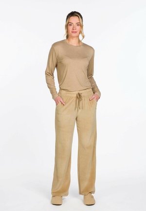 Vrouw staand met handen in de zakken, gekleed in een beige top met lange mouwen, losse beige broek met trekkoord, en beige sloffen, op een witte achtergrond.