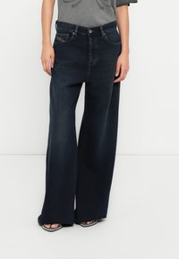 Τζιν παντελόνι με φαρδύ πόδι σε σκούρο τζην ύφασμα και ψηλή μέση, με διακριτική ξεθωριασμένη όψη, εμπρόσθιες τσέπες και κούμπωμα με ασημένιο κουμπί. Λείο υφή.