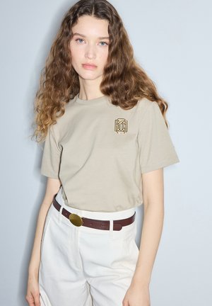 Młoda kobieta z kręconymi brązowymi włosami, ubrana w beżowy t-shirt ze złotym emblematem i białe spodnie z brązowym paskiem na jednolitym tle.