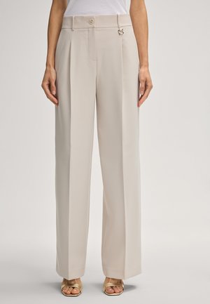 Beige broek met wijde pijpen en plooien aan de voorkant, sluiting met knoop en haak, gecombineerd met gouden sandalen met open teen en een wit topje.